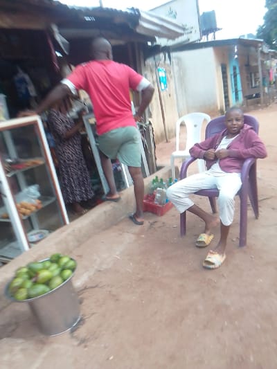 Here is a forensic description of the scene:

**Location:** Owerri, Nigeria. The scene appears to be in a semi-urban or roadside location.

**Subjects:**
*   A young individual is seated in a purple plastic chair wearing light-colored pants, a white shirt, a maroon jacket, and slides.
*   A man in a red shirt and green shorts stands near a shop.
*   Another individual in a long printed dress stands near the shop.

**Objects:**
*   A metal bucket filled with green fruits.
*   A glass-fronted display case exhibiting food items is located at the shop.
*   A red plastic crate.
*   Plastic bottles.
*   Two plastic chairs (purple and white).
*   A building with a corrugated metal roof and various structural elements.
*   A building with a corrugated metal roof and a water tank on top of it.

**Actions and Interactions:**
*   The seated person is positioned in a chair in front of the shop, facing the camera.
*   The man in the red shirt appears to be interacting or associated with the shop.

**Environment:**
*   The ground is a bare earth surface.
*   The background includes a mix of building structures and what appears to be a street or roadside environment.
*   Evidence of a small commercial establishment/shop is seen.