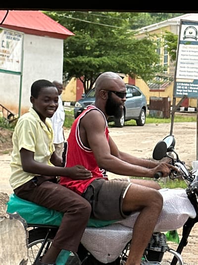 Two individuals are seated on a motorcycle in Mararaba, Nigeria. The person in front, a young male, wears a light yellow short-sleeved shirt and brown trousers. He is smiling and looking to his right. The person behind him, an adult male with a beard and sunglasses, wears a red tank top with white trim and dark shorts. He is looking forward, operating the motorcycle. The motorcycle itself has a green seat cover and a white bubble wrap-covered parcel behind it. In the background, a light blue car is parked, and there are buildings and trees. A signpost with text is visible to the right, indicating a location likely within Mararaba, Nigeria. The ground appears to be a dirt or unpaved road.