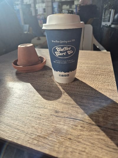 An indoor scene in Hereford, United Kingdom, displays a light brown wooden table surface. On the table, a blue disposable coffee cup with a white lid, prominently featuring "Coffee Cart Co" branding and the text "Hereford's Own Roasted Independent Coffee Roasters," stands upright. To the left of the cup, a small, inverted terracotta pot rests on a terracotta saucer. Direct sunlight from the left illuminates the foreground, creating distinct shadows of the cup and pot on the table. The background is out of focus, revealing blurred shapes consistent with an indoor public space, including indistinct figures and structural elements. No overt actions or interactions are visible.