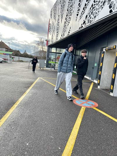 The scene depicts an outdoor paved area adjacent to a modern commercial building in Hœnheim, France, under an overcast sky. Three individuals are present. In the foreground, two young males walk towards the left. The male closer to the camera wears a black beanie, a grey-blue puffer jacket, light grey sweatpants, and black sneakers, carrying a dark backpack. He looks slightly to his right with a subtle smile. The male beside him wears a dark long-sleeved top, dark pants, and dark sneakers, also with a dark backpack; he looks slightly to his left with his mouth open and his left hand slightly raised. In the midground, a third individual, dressed in black, walks further away, looking down at a mobile phone held in their left hand.
The ground is dark asphalt pavement with bright yellow painted lines forming a directional cross in the foreground. A circular red and blue "No Parking" sign is painted on the asphalt, partially obscured by the feet of the two foreground males. To the right, a large, multi-story building features a distinctive metallic gray facade with intricate cut-out patterns and a red vertical stripe. The ground level includes glass automatic doors, through which a "FRAIS" sign is visible, suggesting a supermarket. Dark grey building sections with yellow and black striped safety barriers are also present. In the background, to the far left, a white car, green structures, and a house with a reddish-brown roof are visible. A line of leafless trees spans the distant horizon beneath a heavy, cloudy sky.