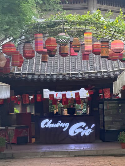 This is a daytime, eye-level, outdoor shot of what appears to be a market or cafe stall with a thatched roof and traditional Vietnamese lanterns hanging overhead. The lanterns are cylindrical and have intricate, colorful patterns. Below the roofline, small Vietnamese flags are strung together, indicating a potential patriotic or festive atmosphere. A counter with a glowing white sign that reads "Chuông Gió" is visible, suggesting the establishment's name. The flooring is paved with bricks. The environment is green, with trees and foliage visible in the background, hinting at a possible park or natural setting within the city of Tây Hoa Lư Ward, Vietnam.