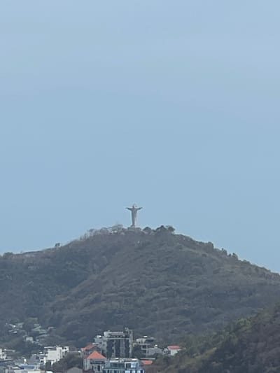 The image shows Christ the Redeemer statue atop Corcovado Mountain. A vast, light blue sky contrasts with the textured, verdant mountain. Buildings dot the lower right edge, suggesting an urban setting. The statue, a light gray, stands prominently against the mountainside, its arms outstretched.