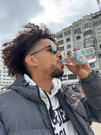 A male subject is depicted in a mid-shot, viewed in profile facing right, actively drinking from a clear plastic bottle of Ain Saïss natural mineral water. He possesses dark, curly hair styled with a fade, a short beard, and wears dark, circular-frame sunglasses. His attire consists of a gray puffer jacket worn over a light gray hooded sweatshirt, which features a dark blue drawstring with tassels. The background reveals an urban setting under an overcast, light gray sky. A prominent multi-story concrete building, appearing aged with numerous windows and balconies, occupies the space behind the subject. Additional buildings are visible further in the background to the left. The scene is located within an urban area of Tangier, Morocco, characterized by dense architectural structures typical of a city environment.