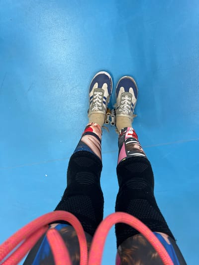 The image shows a person's legs from a top-down perspective. The person is wearing blue and white sneakers, printed socks with images of faces, black knee-high compression socks, and is standing on a light blue floor. A pink rope is draped across the right side of the person's legs. The location is not identifiable as Berezniki, Russia, based on the visual information.