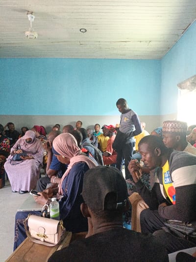 An indoor scene in Fagge C, Nigeria, depicts a gathering of approximately 20-30 individuals within a room featuring light blue walls above a grey wainscoting and a white paneled ceiling with a visible ceiling fan and a recessed light fixture. Natural light enters from a bright window on the right.
In the foreground, a person with their back to the camera, wearing a black baseball cap and t-shirt, is seated. To their right, another seated male wears a yellow t-shirt with a colorful graphic under a striped grey and black long-sleeved top; he holds a small, light-colored object in his right hand. A standing male, wearing a grey and blue long-sleeved shirt with a logo, is positioned slightly right of center, looking downwards.
Seated in the mid-ground, a woman wearing a light pink hijab and dark blue dress is focused on a white laptop and smartphone resting on a wooden surface in front of her. A clear plastic water bottle and a light pink handbag with a gold clasp are also on this surface. To her left, another woman in a light purple hijab and full-length dress, with a black face covering, looks down at an object in her lap.
Additional men and women are seated in rows behind these figures, extending into the background, some wearing traditional head coverings. The overall scene suggests a community meeting or gathering.