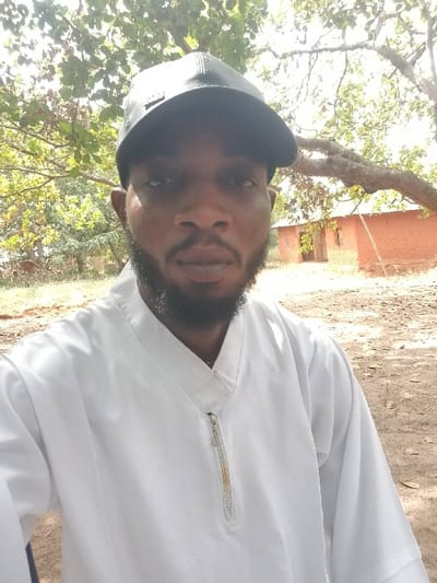A male individual is depicted in a close-up, chest-up outdoor shot, identified as being in Ochadam, Nigeria. He is an adult male with dark skin, a beard, and a mustache. He wears a black baseball cap and a long-sleeved white garment featuring a V-neck collar and a vertical zipper closure extending from the collar to the upper chest, which exhibits minor discoloration. The individual faces forward, looking directly into the camera. The background consists of sparse to moderate green foliage from multiple trees overhead and in the mid-ground. To the right, two single-story structures constructed from reddish-brown material, possibly earth or brick, with light-colored roofs, are visible under the branches of a large tree. The ground is dry, light brown earth.