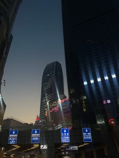 This media file captures a modern cityscape at dusk or early night in Liangjiang, China, dominated by illuminated buildings against a dark blue sky that brightens towards the horizon.
In the center, a tall, dark, curved skyscraper features prominent vertical and horizontal light strips, along with pink heart-shaped decorations or lights on its side, and the text "pro ulo". Below it, on a lower building, a large illuminated sign displays "Dopamine Park" with Chinese text detailing "亲子同乐 解压释放 趣味活动 全龄畅玩" (family fun, stress relief, fun activities, all ages play).
To the top left, another large skyscraper displays bright, vertical Chinese characters "聚星传媒" (Juxing Media). Nearby, smaller illuminated signs read "经络 常楽 推拿 足疗" (Meridian Massage, Health & Longevity, Tuina, Foot Reflexology).
Multiple blue directional road signs with white text are visible, providing navigation cues in both Chinese and English. These include signs for:
- "建北一支路" (Jianbei Ind Branch Rd.) leading to "新上海大厦" (New Shanghai Building).
- "兴塔路" (Xingta Rd.) leading to "CIC天和国际" (CONCORD INTERNATIONAL CENTRE) and "观音桥1号" (THE GRAND ONE).
- "观音桥都市旅游区" (Guanyingiao Urban Tourism Area) leading to "融恒·盈嘉中心" (STAGE), "大融城" (MIX PARK), and "未来国际" (FUTURE INTERNATIONAL).
Several "P" signs indicate parking, specifically "P 未来国际" and "P 天和国际". Other visible text includes "天和里" (Tian He Li) and "ojncord". A dark, textured structure, possibly a building facade or roof, is visible in the lower left. The scene portrays a vibrant urban environment with no people visible.