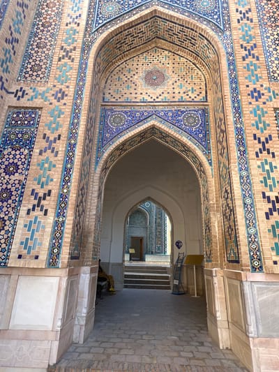 The image shows a detailed view of the entrance to a historical building, likely a madrasah or mosque, in Samarkand, Uzbekistan. The structure features intricate geometric tilework in shades of blue, teal, and beige, set against a brick facade. The archway leads into a courtyard with more tiled architecture visible in the distance. In the foreground, a person is seated on a bench to the left, partially obscured by the wall. To the right, a metal stand with informational signs and a blue ladder-like structure is present. The ground is paved with bricks, and the overall impression is one of rich cultural heritage and architectural splendor.
