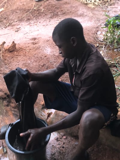 An individual, likely a young man, is seen outdoors in Ukwu-Abwa, Nigeria. He is kneeling beside a black bucket and appears to be rinsing or cleaning a dark-colored cloth within it. The ground is earthen, and there's some vegetation and water visible in the background. The individual is wearing a dark-colored shirt and dark shorts.
