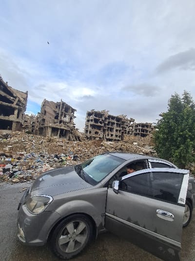 A gray four-door sedan is parked on a wet, dark surface in Joubar Municipality, Syria. The vehicle's driver's side front door is open. A male individual with dark hair is visible in the driver's seat, looking towards the right side of the frame. In the background, a vast expanse of debris and rubble extends, leading to multiple heavily damaged concrete multi-story buildings. These structures show significant destruction, with exposed rebar and fractured facades. A solitary green tree stands on the right side of the image, behind the car and partially obscured by the rubble. The sky is overcast with a mix of light and dark clouds, and a single dark bird is captured in flight near the top center of the frame.