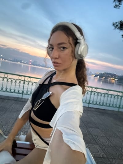 A young woman with curly brown hair, wearing white headphones and a black bikini top with a white shirt draped over her shoulders, takes a selfie. She is outdoors near a body of water, likely a river or lake, with a city skyline visible in the distance. The sky is overcast with hints of pink and orange, suggesting either sunrise or sunset. She is seated on a bench with a decorative metal railing behind her and a patterned paved walkway. Sunglasses are clipped to her bikini top. The overall impression is a casual, possibly post-activity moment, with the woman appearing relaxed and enjoying the view. The setting seems to be a public area with a scenic backdrop, typical of waterfront promenades.