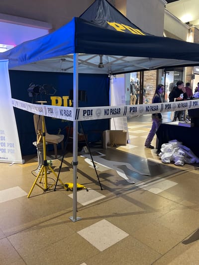 This is a full, eye-level shot of a crime scene setup under a navy blue pop-up tent with yellow "PDI" lettering. The tent is located outdoors, possibly in a public area or plaza, as indicated by the tiled floor and building facades visible in the background, suggesting an urban environment in Arica, Chile.

The scene is cordoned off with white tape bearing the text "PDI NO PASAR," which translates to "PDI Do Not Enter." The tape stretches across the foreground, partially obscuring the view of what is beyond.

Inside the tent, several elements suggest a simulated crime scene. A yellow tripod with two spotlights is positioned on the left, aimed towards the back of the tent. Behind the spotlights, a dark blue banner with large yellow "PDI" lettering is visible. A simple chair covered with a light-colored fabric sits in the middle ground. On a pedestal to the left of the chair, a small, decorative vase is placed. In the foreground, on the floor, there is a white silhouette outline of a human body, indicating a simulated victim. Scattered white papers or fabric are piled on the right side of the tent.

In the background, through the open sides of the tent, glimpses of shops and pedestrians are visible, suggesting the scene is set up in a public or commercial area. Two individuals are partially visible on the right side of the frame, near the edge of the tent, observing the scene or interacting with personnel. One appears to be a woman with dark hair, and the other is a person with their back to the camera. The lighting is artificial, with blue and yellow hues contributing to the overall atmosphere.