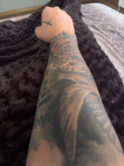 The image displays a close-up view of a person's forearm and hand, adorned with extensive tattoos. The arm is positioned on a dark, textured blanket or rug. On the back of the hand, near the knuckles, a small black "X" tattoo is visible. The forearm features several intricate tattoo designs, including what appears to be a clock face with a red rose, and other abstract or pictorial elements in shades of black, grey, and red. Some text is also visible as part of the tattoo art. The background consists of a bed with a patterned sheet. The lighting suggests an indoor setting. No specific location in Bansko, Bulgaria, can be determined from the visual information provided in this image.