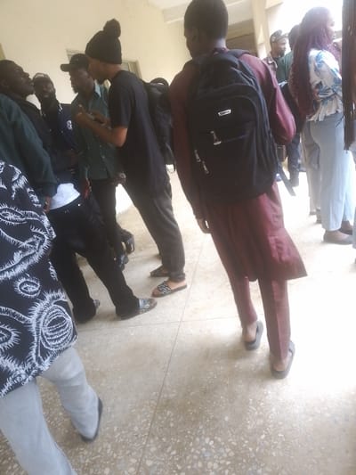The image depicts a group of young adults gathered in an outdoor, possibly campus, setting. The individuals are dressed in casual attire, including a person wearing a maroon outfit and a backpack, another in a black t-shirt and black pants, and others in shirts and trousers. Some are wearing hats, and one has a distinctive black beanie with a pom-pom. The ground appears to be a paved area with scattered stones. The lighting suggests it is daytime, possibly with bright sunlight creating some overexposed areas. The scene captures a moment of informal gathering or interaction among the individuals, with some looking at their phones. The environment suggests a location in Rafin Par, Nigeria, given the context.