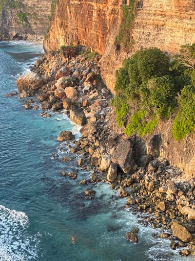 An aerial view of a rocky coastline with a high cliff in Pecatu, Indonesia, bordering the ocean. The cliff face on the left side of the image is a mix of brown and orange tones and covered with green vegetation on the top. Below the cliff, there's a rocky area with many large, brown-colored rocks. The water near the rocks is light blue and clear, becoming deeper blue further away from the shore. White foamy waves crash against the rocks, and a person is swimming in the water.
