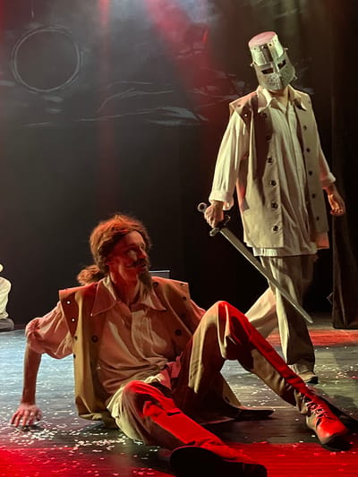 The scene depicts a theatrical performance unfolding on a stage in Novoshakhtinsk, Russia. Two male figures are central to the composition. In the foreground, one figure is seated on the dark stage floor, leaning backward with his left hand supporting him and his legs extended forward. He wears a light-colored long-sleeved shirt, a tan sleeveless vest with dark circular details, dark trousers, and dark lace-up boots. His hair is reddish-brown, tied back, and he sports a prominent mustache and goatee. His head is turned to his right, looking upwards, with an intense expression. Behind and to the right of the seated figure, a second individual stands, facing generally towards the foreground figure. This standing figure wears a light-colored long-sleeved shirt and a tan sleeveless coat-like garment. His head is fully covered by a metal bucket-style helmet, featuring horizontal eye slits. He holds a silver-colored sword with a dark hilt, pointed downwards, in his right hand. The dark stage floor is speckled with small white debris, resembling confetti. The background is a dark theatrical backdrop adorned with abstract white brushstroke-like markings, including a distinct large white circle in the upper left portion. The scene is illuminated by dramatic stage lighting, with notable red and white spotlights.