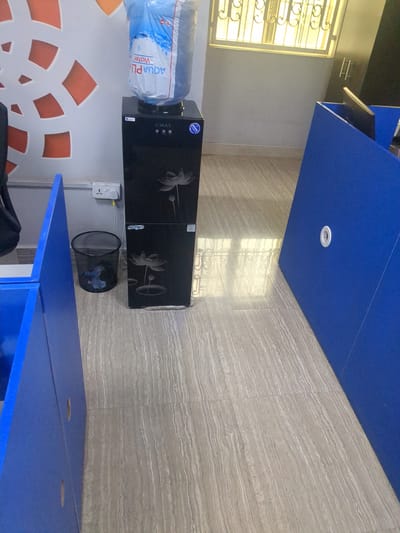 The image depicts a water cooler with a blue water jug on top, situated in what appears to be an office or communal space. The water cooler is black with decorative white lotus flower motifs. Next to the water cooler is a black mesh trash can containing some blue and black debris. A white electrical outlet with a yellow cord plugged into it is visible on the wall behind the trash can. The floor is tiled with a light brown and gray striped pattern. To the left and right of the scene are blue desk dividers or cubicles, one of which has a circular white grommet. In the background, a window with decorative metal bars and a yellow tint can be seen. The overall impression is of a functional, possibly shared, workspace. The image does not provide any direct or indirect information about the city of Fagge A, Nigeria, or any specific location within it.
