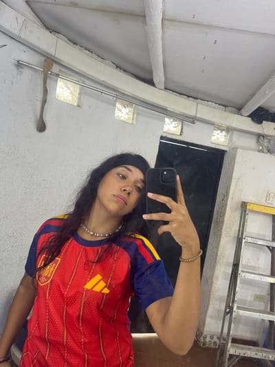 In Porlamar, Venezuela, a young woman with long, dark hair tied back with a black headband, poses for a selfie in a red and blue soccer jersey. She wears a beaded necklace and a bracelet on her right arm. She holds a black smartphone in her right hand, angled to capture her reflection. The background is a rough, white wall with exposed beams and ventilation grates. To the right, a silver ladder with yellow accents leans against the wall. The scene appears to be an indoor workshop or storage area, possibly in a home.
