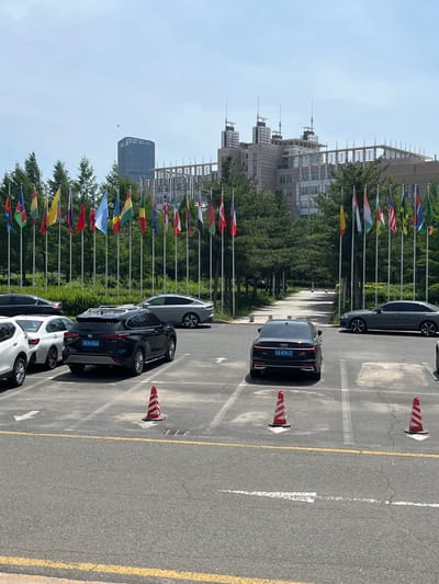 A view of a parking lot in Changchun City, China, taken from an elevated perspective. The parking lot contains multiple vehicles parked in rows. Several safety cones are positioned on the lot's surface. A series of flags, likely representing different nations, are displayed along the edge of the parking area. In the background, buildings and trees are visible, indicating an urban environment. The sky is partly clear. The photograph appears to have been rotated counter-clockwise, shifting the horizontal orientation of the scene.