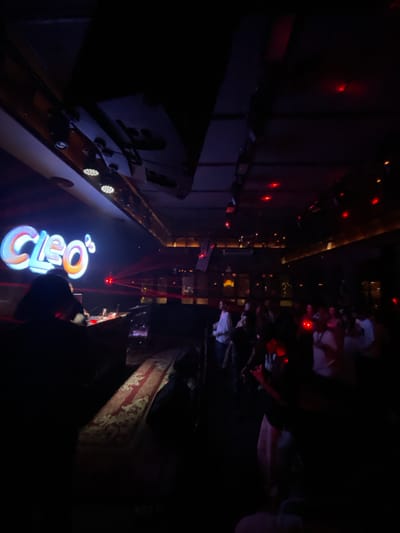 The image depicts an indoor scene, possibly a nightclub or bar, in Central Jakarta, Indonesia. A large, illuminated sign displaying the word "Cleo's" in colorful, stylized lettering is visible on the left side of the frame. Below the sign, a person, likely a DJ, is silhouetted behind a console, with red laser lights projecting across the scene. A crowd of people, many of them indistinct due to the low lighting and focus, are gathered in the dimly lit space, suggesting a lively atmosphere with music and dancing. Red ambient lighting is present throughout the venue, casting a red glow on some of the patrons. A patterned rug is visible on the floor in the foreground.