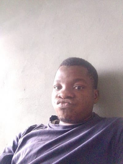 This is a selfie of a young Black man, likely taken indoors against a plain, light-colored wall. The lighting suggests it could be daytime, possibly with diffused natural light, as there is a shadow cast on the wall to the right of the subject. The man is wearing a dark-colored long-sleeved shirt. He is looking directly at the camera with his lips pursed, giving a playful or perhaps slightly sulky expression. His short, dark hair is visible. The background is a simple, textured wall, offering no specific environmental clues beyond being an indoor setting. There is no visible text in the image.