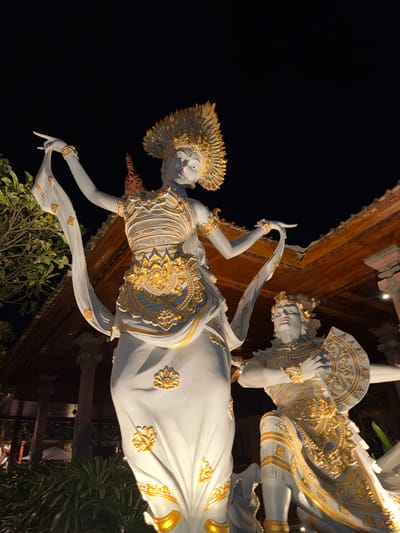 This media file features two statues in a nighttime setting, likely in Kuta, Indonesia. The statues depict figures in traditional Balinese attire, characterized by elaborate gold ornamentation over white fabric. The main subject is a female figure in the foreground, adorned with a headdress, intricate bodice, and flowing garments. Her pose suggests a graceful dance or ritual, with one arm extended and her hand delicately held. Behind her, another figure, also in ornate golden attire, is visible, its head tilted and hand raised, potentially mirroring the first figure's movements or engaged in a complementary gesture.
The statues are illuminated, casting shadows and highlighting the detailed craftsmanship of the gold accents against the white material. The background is a dark night sky, with glimpses of a wooden structure overhead, possibly part of a temple or a decorative archway. Lush green foliage is visible at the bottom, adding a natural element to the scene. The overall impression is one of artistic elegance and cultural significance, evoking a sense of ancient traditions and spiritual performance. There is no visible text in the media file. The time of day is clearly night, indicated by the dark sky and artificial lighting.