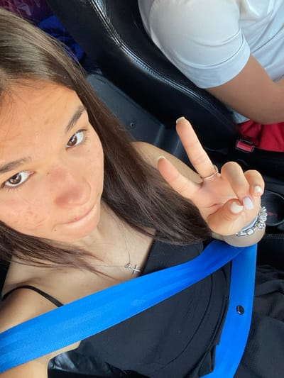 This is a close-up, high-angle, point-of-view photograph of a young person in a car. The person, who appears to be female, is wearing a black top and a silver necklace with a cross pendant. A bright blue seatbelt is prominently displayed across the frame. The person is making a peace sign with their right hand, which is adorned with rings and a charm bracelet. Their nails are painted white. The background includes the black interior of a car, with a glimpse of another person's arm and shoulder on the left, wearing a white shirt. The image appears to be taken during daylight. There is no visible text in the image. The setting is likely a car in Sevlievo, Bulgaria. The person's face is partially visible, and they appear to be looking up, possibly at the camera. Their skin shows some texture and a few small blemishes.