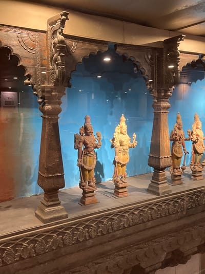 This image depicts a collection of ancient Indian wooden sculptures displayed behind a glass enclosure. The sculptures appear to be religious figures, possibly deities, adorned with traditional clothing and jewelry. They stand on pedestals, arranged in a row within a museum setting. The backdrop is a vibrant blue wall, creating a striking contrast with the warm tones of the wooden carvings. The architectural elements in the foreground and above the sculptures suggest an ornate wooden frame, intricately carved with decorative patterns and motifs. These elements hint at traditional Indian temple architecture or craftsmanship. The overall scene is well-lit, highlighting the details of the sculptures and the surrounding artifacts. This scene is likely part of a museum in Mumbai, India, such as the Chhatrapati Shivaji Maharaj Vastu Sangrahalaya (CSMVS), known for its extensive collection of Indian art and artifacts.