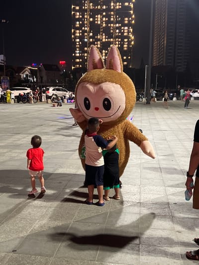 This nighttime image captures a lively urban public square, identified as the plaza at Vinhomes Central Park, adjacent to Landmark 81, in the Binh Thanh District of Ho Chi Minh City, Vietnam.
In the center, a person in a large, brown, furry mascot costume, resembling a monkey or rabbit with a light pink face, large dark eyes, and a wide smile, stands with its left hand raised in a waving gesture. The mascot wears gold-colored bead necklaces. Two young children, seen from behind, are embracing the mascot at its midsection. The child on the left wears a light-colored shirt with dark sleeves and dark shorts; the child on the right wears a dark shirt and shorts. To their left, a third young child in a red shirt and light shorts stands with their back to the camera, facing the mascot. Part of an adult holding a light blue bottle is visible on the far right foreground.
The ground consists of large, light gray square and rectangular paving tiles, showing multiple elongated shadows from the subjects and ambient lighting.
The background on the left features several parked motorcycles and cars, with groups of people gathered around. Beyond them, a row of low-rise buildings with illuminated windows is visible, along with a partial red neon sign on a distant building. The background on the right is dominated by a very tall, dark skyscraper, clearly identifiable as Landmark 81, with numerous brightly lit windows across its facade, indicating residential or office occupancy. An adjacent, less illuminated high-rise is also present. The scene is artificially lit, creating distinct light and shadow patterns.
