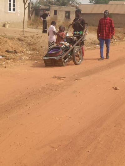 A sandy path leads to a group of people. One man wears a red plaid shirt and blue pants, walking toward the camera. Two children are with a man pushing a cart filled with assorted items, including tires and plastic containers. Another person is standing in the background, dressed in dark clothing. Buildings are visible in the background. The scene is sunlit, creating shadows on the sandy ground.