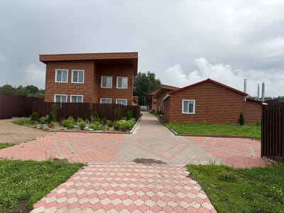 The image shows a street with two wooden houses on a cloudy day. The houses are modern with large windows and dark brown wood siding. The street is paved with pink and white interlocking pavers, and there is grass on either side of the street. The houses are surrounded by a wooden fence. The location is the city of Пакли, Russia.