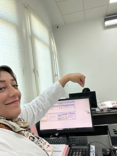 A woman wearing a white coat is posing for a selfie in what appears to be a medical office. Behind her is a computer monitor displaying lines of data. To the monitor's right is a calculator and a keyboard. The background includes a window with white blinds. A security camera is visible on the ceiling. The woman is wearing bracelets and has her hand raised near the monitor.