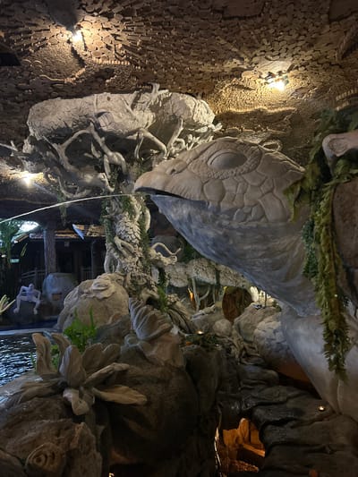 The image depicts a large, sculpted dragon head and a body with intricate carvings of leaves and branches, located at the Bali Pulina Agro-Tourism plantation in Mengwi, Indonesia. The dragon sculpture is part of a decorative water feature, with a stream of water emerging from its mouth and flowing into a pool below. The surrounding environment features rock formations, lush greenery, and a rustic wooden building with a tiled roof. The ceiling is adorned with wooden logs, and ambient lighting creates a mystical atmosphere. In the foreground, a stone sculpture of a flower rests on a rock. Other decorative elements include a frog-like sculpture near the water and a distant view of the plantation's structures.