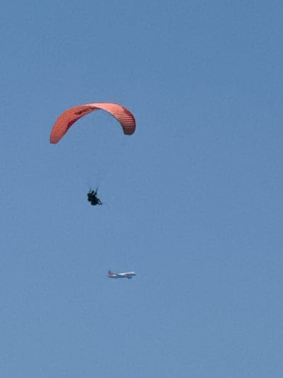 An orange and red paraglider is suspended in the clear blue sky above Boreti, Montenegro, with a person seated beneath it. Below the paraglider, a small white and red airplane is also visible, flying in the same direction. The scene depicts a recreational activity in an open, outdoor environment.