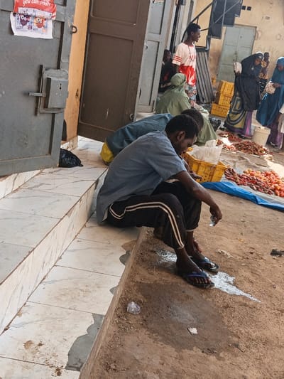 An outdoor market scene is depicted in Kawaji, Nigeria, under bright daylight. In the immediate foreground, a tiled step transitions to uneven, dry dirt ground. A white liquid substance is spilled on the dirt, along with a discarded clear plastic wrapper.
A man is seated on the tiled steps in the mid-left, facing towards the right. He wears a light blue short-sleeved shirt, black track pants with two white vertical stripes, and blue flip-flops. He holds a small, clear, cylindrical object in his right hand. Directly behind him, another individual is partially visible, seated, wearing a teal-green garment, with their back to the viewer.
To the left, a large, dark grey metal door with a robust lock mechanism is visible, partially open. A red and white poster displaying text and illustrations is affixed to the upper left portion of this door. Another partially open dark door is seen further back.
In the mid-right and background, several individuals are present, suggesting a bustling market environment. A man stands, facing slightly left, wearing a white short-sleeved top with red markings and a patterned lower garment. He has short dark hair. To his right, multiple women, primarily in traditional Nigerian attire including head coverings, are seen. One woman in a dark abaya stands near piles of produce.
The ground to the right of the seated man features a blue tarpaulin laden with numerous red fruits, identified as tomatoes and peppers. Several yellow plastic crates are interspersed among the produce and people, indicating commercial activity. The background consists of light-colored building walls, some appearing weathered.