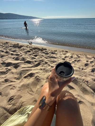 A first-person perspective shot captures a serene moment on the sandy beach of Nesebar, Bulgaria, with the Black Sea shimmering under a clear blue sky. In the foreground, a person's tanned legs are visible, one hand holding a clear travel mug with a black lid. A gold-strapped wristwatch is worn on the left wrist. The beach sand is textured with footprints and ripples. In the mid-ground, a seagull stands alert, facing the camera. Further out in the water, a lone figure walks into the sea, their silhouette stark against the sun-drenched surface. The background features the hazy outline of a distant coastline with rolling hills under the bright sun.