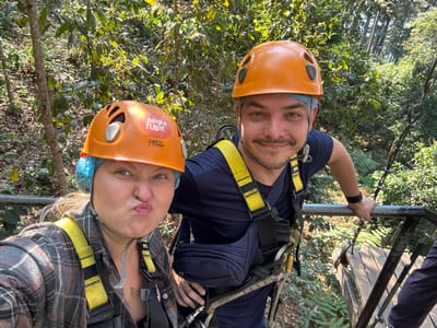 Two individuals, one female and one male, are depicted on an elevated platform in a dense jungle environment within the city of Pa Miang, Thailand, consistent with a zipline or canopy adventure activity. Both wear orange Petzl climbing helmets over blue hairnets and yellow full-body safety harnesses. The female, positioned closer to the camera on the left, has light-colored hair and is wearing a plaid shirt in shades of brown, grey, and white. She is making a puckered, pouting facial expression directly at the camera. The male, positioned to the right, has dark hair and stubble, and is wearing a dark blue short-sleeved shirt. He is smiling towards the camera. Both individuals are partially grasping a silver metal railing in the foreground. Behind them, lush green foliage dominates the background, with further wooden and metal platform structures visible to the right, descending into the forest. The female's helmet features a "Jungle Flight" sticker.