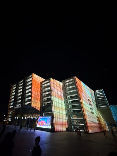 The image captures a nighttime scene in Jiangbeicheng Subdistrict, China, with a large, modern building illuminated by colorful lights. The building features a facade with a grid pattern, covered in projected colors ranging from orange and yellow to blue and green, creating a vibrant, abstract design. In front of the building, a large screen displays an animated scene with cartoon characters. Several people are visible in the foreground and mid-ground, some walking and others standing, appearing to be observing the building or the screen. The sky is dark, indicating it is nighttime, with faint lights and what appears to be a distant satellite or celestial body. The ground is paved, and the overall atmosphere suggests a public gathering or a visually striking urban display.