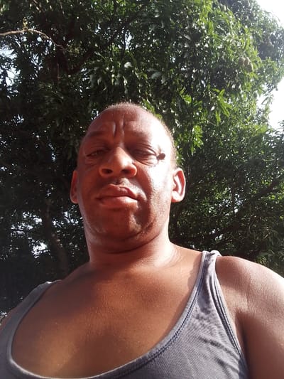 An outdoor, eye-level photograph depicts a dark-skinned adult male in Narayi, Nigeria. The subject, appearing middle-aged with a closely cropped or bald scalp, is positioned centrally and faces the camera directly. His expression is neutral, and his eyes are slightly narrowed, indicative of bright ambient light. He is clad in a grey ribbed tank top. A visible sheen is present on his forehead and exposed upper torso. The background consists of dense green arboreal foliage, with a bright sky visible through gaps. Small, spherical, light-green fruits are discernible among the leaves in the upper right, consistent with the produce of a fruit-bearing tree. The scene is illuminated by strong overhead or frontal sunlight.