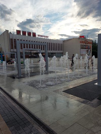 A photograph taken in Zheleznogorsk, Russia, shows a public square with a water fountain display. The fountain features numerous jets of water spraying upwards from the ground, creating a dynamic visual effect on the wet pavement. Several children and adults are present in the scene. One boy is riding a bicycle, while another individual stands near him. In the background, a building with a prominent sign reading "КИНОТЕАТР" (Cinema) and "РУСЬ" is visible. Another sign above the entrance advertises "КАФЕ ХРУС ТАЙМ" (Cafe Krus Time) and "ДОСТАВКА" (Delivery), along with a digital clock displaying "24:06". The sky is overcast with clouds. The scene suggests a leisure activity in a public space.