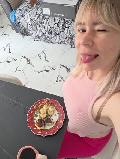 The image appears to be a selfie taken indoors, possibly in a home or apartment. A fair-skinned individual with blonde hair, wearing a pink top and red leggings, is positioned in the foreground with their tongue sticking out.

A black table to the upper-left holds a cup of coffee and a plate. The plate is decorated and contains what appears to be sliced bananas, nuts, seeds, and a browned food item, possibly pancakes.

The background features white tile flooring with black veining. In the upper right, a decorative pillow and a box-like object are visible.

Without specific location details, it is not possible to verify if this is indeed in Batumi, Georgia, but it's an indoor scene with food, personal items, and flooring consistent with modern living spaces.