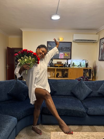 The image depicts an indoor scene, likely a living room, in Alaguntan, Nigeria. An adult female, appearing to be of African descent with short dark hair, stands prominently in the center. She wears an oversized white collared shirt, functioning as a dress, and has red nail polish on her fingers and toes. A silver-colored ankle bracelet is visible on her left ankle. She is smiling and looking towards the camera, with her left leg bent and her right leg extended forward, foot flat on the ground. Her left arm holds a large bouquet of red roses with green foliage and white filler flowers, while her right arm is raised and bent at the elbow.
The background features a dark blue sectional sofa with matching throw pillows on the left and mid-ground. The floor is covered with a light brown carpet, and a light-colored rectangular coffee table is partially visible in the foreground. The walls are cream-colored with white crown molding. On the left, a dark wooden door is partially ajar. Along the back wall, a wooden console table or cabinet is adorned with various objects: several dark figurines depicting human and animal forms (including a giraffe), a small fish tank containing fish, a brown box, some white items, a pair of dark boots, and a statue resembling a meditating figure. To the far right, dried decorative branches are in a vase. Above the cabinet, a large dark-framed painting of a cityscape is centrally mounted, flanked by a yellow emergency exit sign above it and two smaller colorful cards below it on the wall. To the right of the painting, a white wall-mounted air conditioning unit is installed, and further right, another dark-framed abstract artwork with red accents is visible. The white ceiling features a single circular flush-mount LED light fixture.
