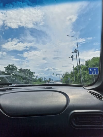 The image shows a view from inside a vehicle looking through the windshield at a road in Zmeyka, Russia. The sky is blue with scattered white clouds. There are trees lining the road, and several lampposts are visible. Traffic lights and road signs are also present. The dashboard of the car is in the foreground.