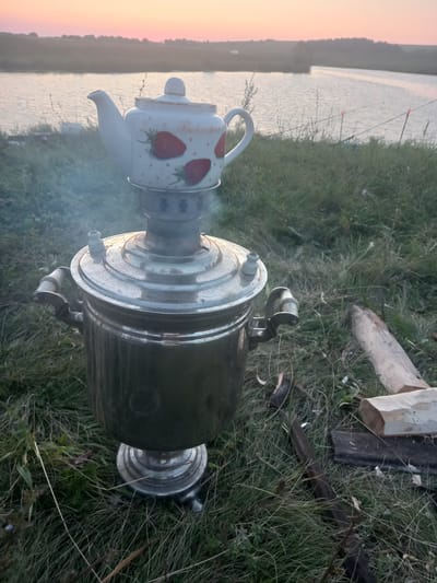 A samovar with a white teapot decorated with strawberries sits on a grassy bank near a body of water. The sky is painted with hues of pink and orange, indicating either sunrise or sunset. Several fishing rods are set up along the water's edge. Some firewood is scattered on the ground next to the samovar. The scene is set in the village of Polevye Ineli, Russia.