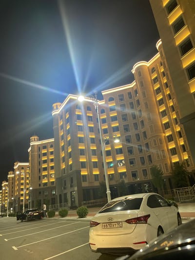 This media file depicts a nighttime scene in a modern urban environment, likely in Grozny, Russia, as suggested by the license plate. The primary subjects are several multi-story buildings illuminated with warm, golden exterior lighting, highlighting their architectural details. The buildings feature numerous windows and decorative elements, contributing to an elegant aesthetic.

In the foreground, a white car is parked, with its rear visible. The license plate on the car reads "B 610 XT 95 RUSSIA". A portion of another dark-colored car is also visible, parked adjacent to the white car. A single person is standing near the dark car, seemingly engaged in an activity related to the vehicle.

The scene is lit by artificial lights from the buildings and streetlights, casting bright beams and creating a luminous atmosphere. The sky is dark, indicating that it is nighttime. The presence of well-maintained buildings, paved parking areas with clear markings, and manicured green bushes suggest a planned urban development or a residential complex. The overall impression is one of a well-lit and orderly urban space at night.