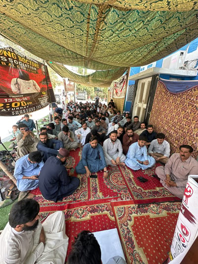 In Quetta City Tehsil, Pakistan, a gathering of individuals, primarily men, are seated on a red carpet under a patterned canopy. The scene appears to be an outdoor assembly. The individuals are dressed in traditional Pakistani attire, with many wearing shalwar kameez. Some are engaged in conversation, while others are looking at their phones or listening intently. 

The canopy is made of a green and gold patterned fabric, creating a shaded area for the attendees. In the background, there are buildings, including one with a blue facade and an air conditioning unit. A banner with text and an image is visible on the left, indicating a "Young Doctors Association Balochistan" meeting scheduled for "Tomorrow 13-06-26" at "2:00 PM". Shoes are piled up on the left side of the image. The overall atmosphere suggests a community or professional gathering.