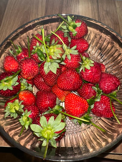 A bowl of fresh strawberries sits on a wooden surface. The strawberries are red, varying in size, and have green stems and leaves attached. The bowl is made of clear glass with a decorative, textured pattern. The image is taken from an overhead perspective. The strawberries are located in a place which might be in Esenyurt, Turkey.