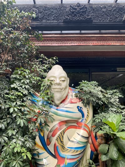 A statue of a figure with a bald head and a serene expression is partially obscured by lush green foliage. The statue is vibrantly colored with swirling patterns of red, blue, yellow, and white. Behind the statue, a building features ornate, dark carved wooden trim above red and dark-banded architectural elements. A glass paneled door or window is visible in the background with a "RECEPT" sign. The scene appears to be an outdoor or semi-outdoor space, possibly a courtyard or garden area within Kathmandu Metropolitan City, Nepal.
