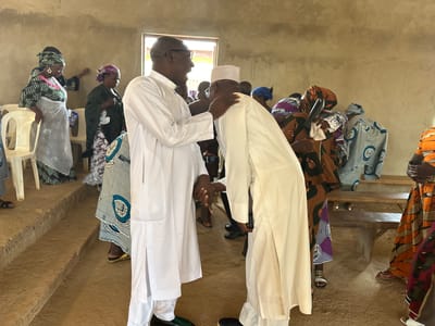 The image captures an indoor gathering in Kufana, Nigeria, possibly a community event or religious ceremony. Two men in traditional white attire are at the forefront, engaged in an embrace, one with his arm on the other's shoulder. The man on the left wears glasses and a white tunic with a high collar, while the man on the right is also in white, including a white skullcap. Behind them, several women are present, some wearing colorful headscarves and patterned dresses. One woman in the foreground wears a green and black patterned dress and headscarf, holding a white cloth. Other women are seen in various traditional garments, some standing and others seated on wooden benches. The environment appears to be a room with unfinished concrete walls and a dirt floor. The lighting suggests it's daytime, with natural light coming from a window visible in the background. Chairs and wooden benches are scattered throughout the space, indicating a gathering of people.