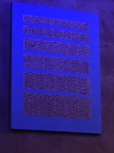 An abstract painting with a bright blue background features six horizontal rectangular sections filled with a textured, dark purple pattern. The pattern in each section consists of repeating, wavy, parallel lines that create a somewhat organic or naturalistic appearance, possibly mimicking wood grain or ripples. The painting is displayed on a wall, which appears to be a dark, textured surface. The overall lighting suggests a dimly lit environment, possibly with a blueish hue casting onto the scene. The painting's location in Mũi Né, Vietnam is not evident from the visual information provided.
